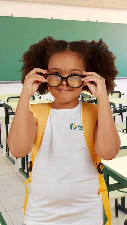 Menina de óculos e mochila amarela vestindo camiseta escolar branca VestCo em sala de aula